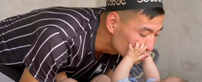 A young father is kneeling on the floor close to his infant as his infant reaches up with his hands to play with his father’s face