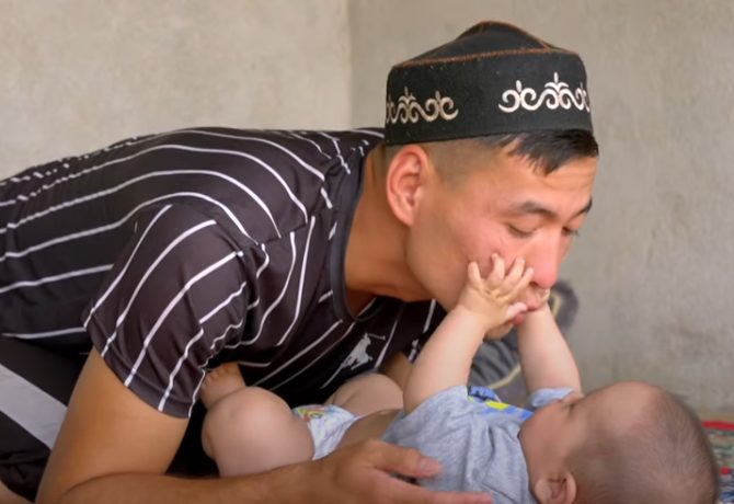 A young father is kneeling on the floor close to his infant as his infant reaches up with his hands to play with his father’s face