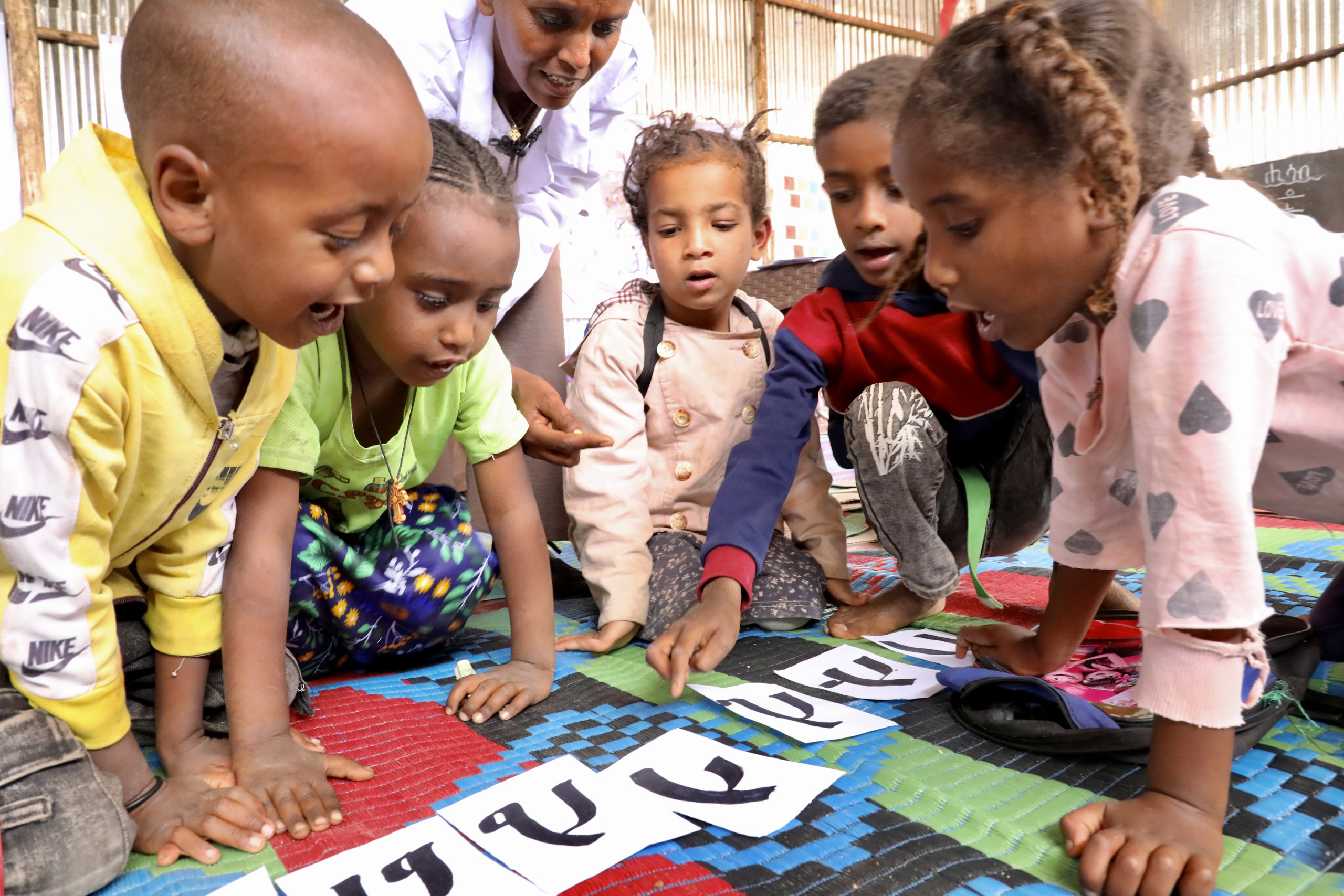 Five children are sitting on a mat pointing at cut-outs of Amharic letters with guidance from a pre-primary educator who is kneeling beside them.