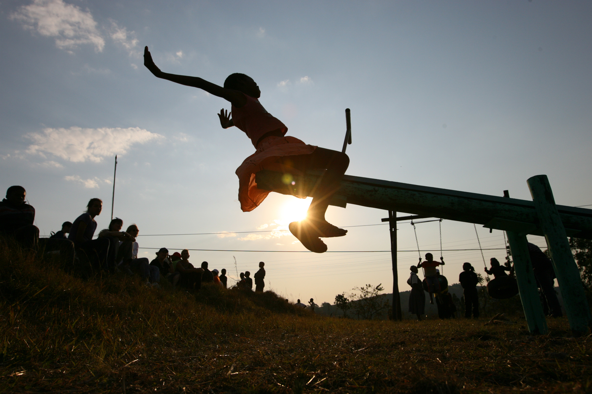 A girl is sitting on a seesaw with her hands up in the air as the sun sets behind her. A group of people are gathered around watching her.