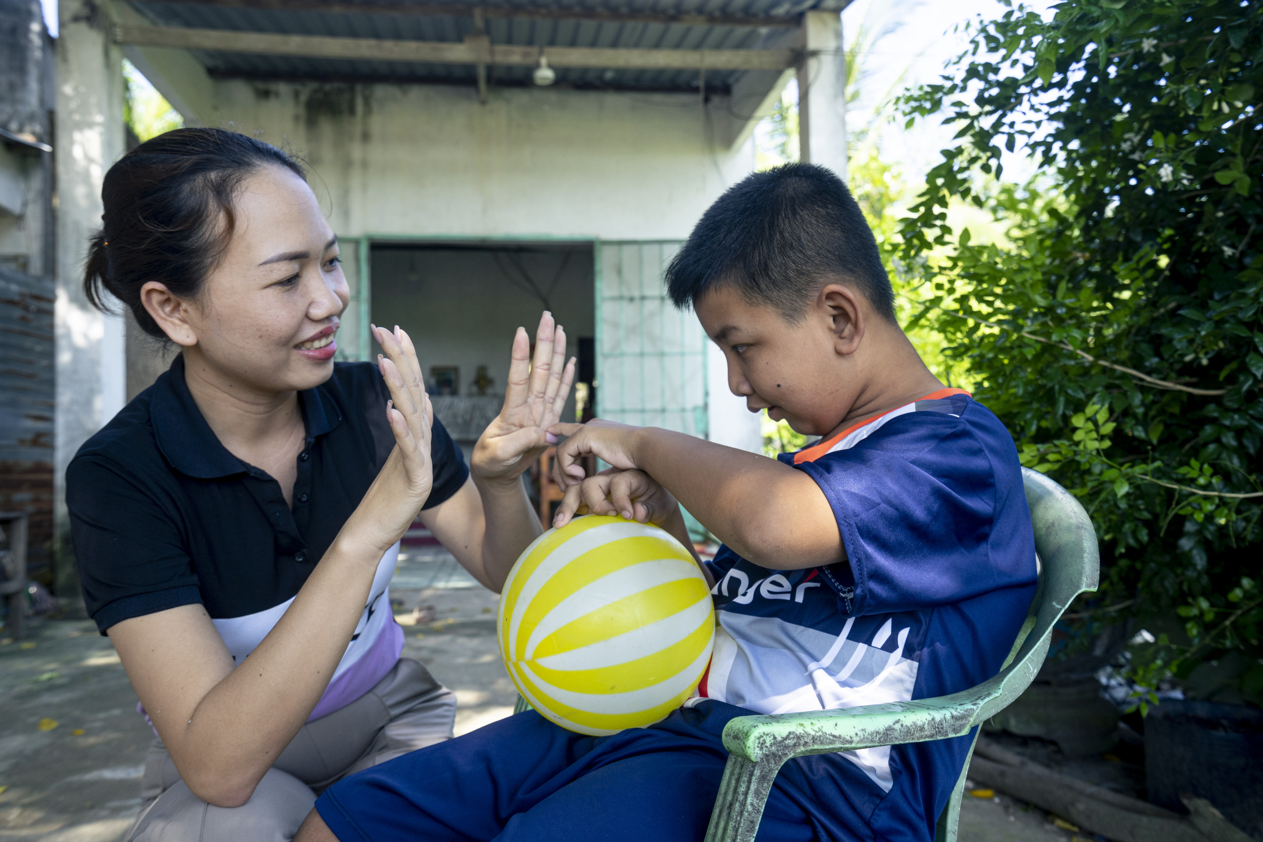 A young boy wearing a blue shirt and shorts is sitting in a green chair pointing at his caregivers fingers while he holds a yellow and white ball. His caregiver is kneeling next to him and smiling.